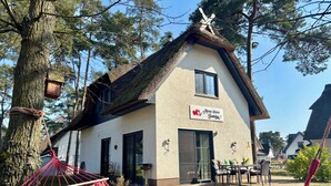 Une maison avec un toit de chaume, une enseigne indiquant « Meine kleine Sanibazar 1 », et un hamac au premier plan.