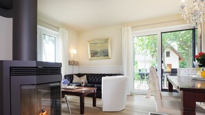 Un salon chaleureux avec une cheminée, un canapé en cuir noir, un fauteuil blanc et une table à manger agrémentée d’un vase de fleurs.