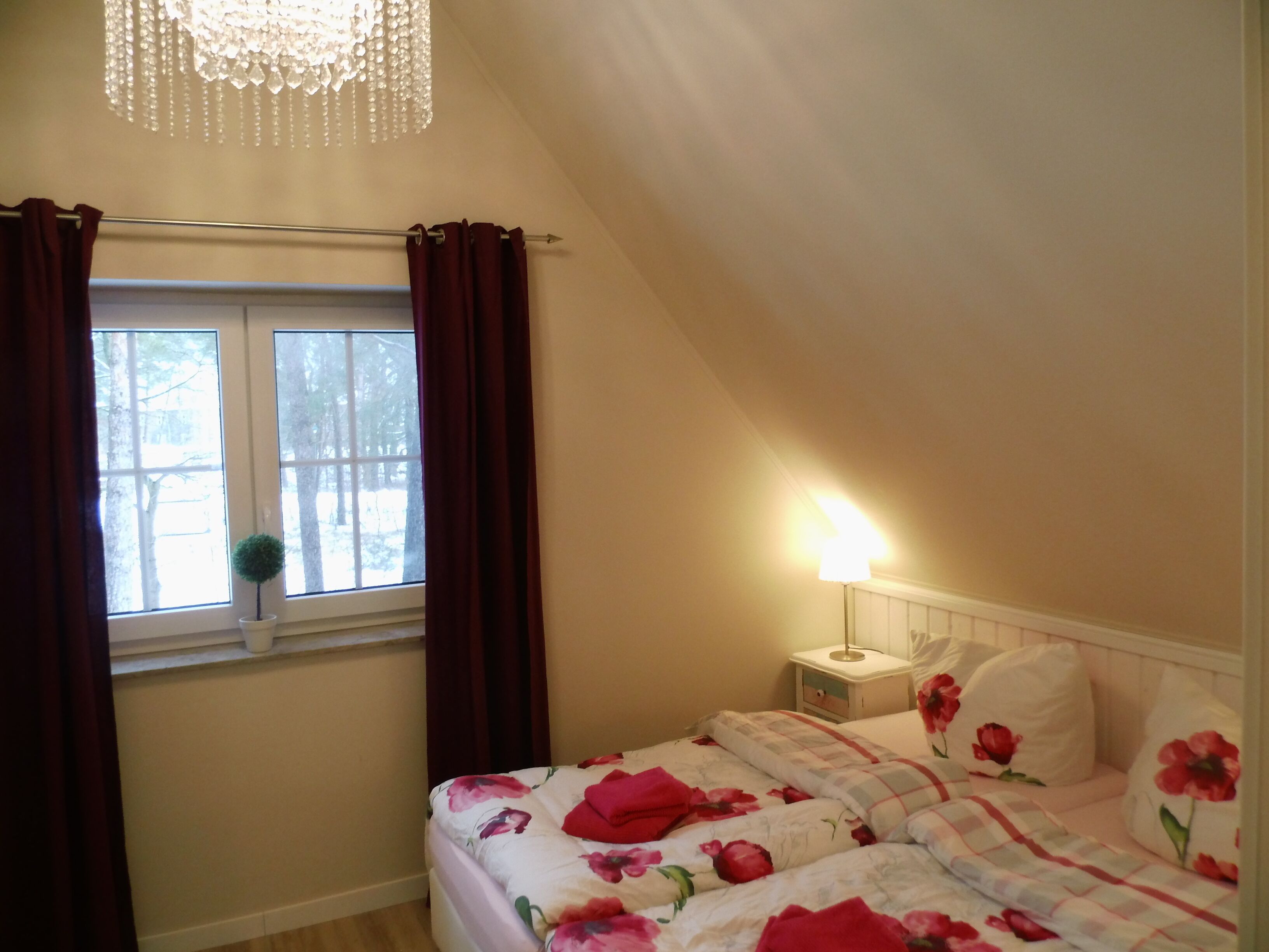 A bedroom with a bed, a window with curtains, a nightstand with a lamp, and a chandelier.