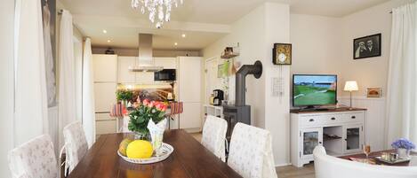 A dining area with a wooden table, white floral chairs, a chandelier, and a kitchen in the background.