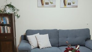 A living room with a blue sofa, two framed floral paintings, a bookshelf, and a coffee table with a vase of flowers. - In the narrow streets of the old town, family-friendly, close to the beach (Ierapetra)