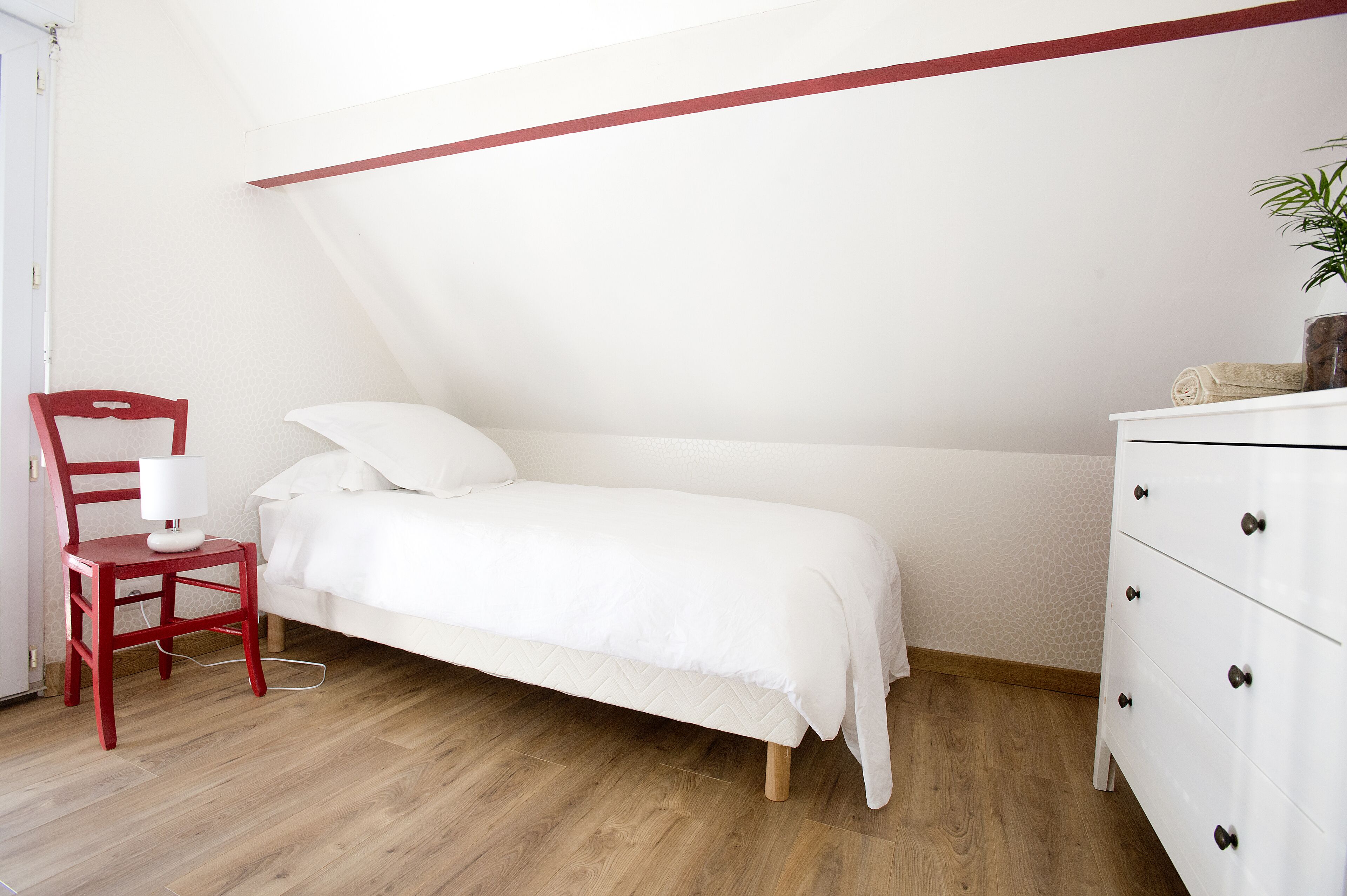 A bedroom with a bed, a red chair, a white dresser, and a plant.