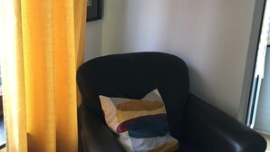 Una poltrona in pelle nera con un cuscino multicolore, un tavolino in legno, una libreria piena di libri e una scatola contenitore blu su una parete bianca.