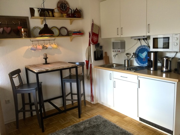 A compact kitchen with white cabinets, a wooden table, two chairs, a microwave, a kettle, and various kitchen utensils.