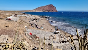 En strand med klippefyldt terræn, et par personer og et bjerg i baggrunden.