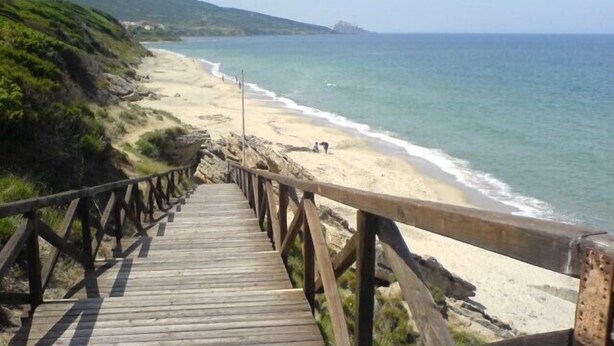Ein Holzsteg führt zu einem Strand mit freiem Blick auf das Meer und sanfte Hügel in der Ferne.