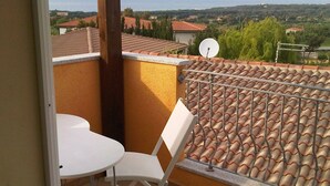 Ein Balkon mit weißem Stuhl und Tisch, einer Satellitenschüssel und Blick auf das Meer und Häuser.