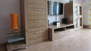 A modern living room with a wooden cabinet, a glass coffee table, a flat-screen TV mounted on the wall, and a small orange vase on the cabinet. - Comfortable holiday home **** (incl. W-Lan) in the best location of Carolinensiel (Carolinensiel)