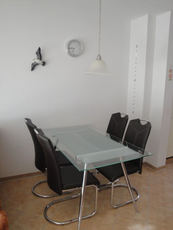 Un coin repas avec une table en verre, des chaises noires, un luminaire suspendu blanc, une horloge murale et un oiseau décoratif.
