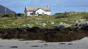 A white house with a grey roof on a grassy hill with rocks.