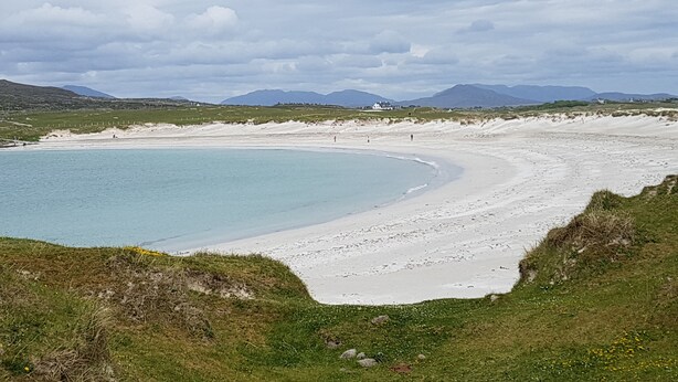A coastal landscape with a sandy beach, a clear blue sea, and distant mountains under a cloudy sky.