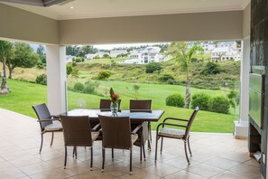 A covered patio with a dining table and chairs, overlooking a lush green landscape and residential buildings. - Modern house, solar-heated pool & stunning views, at Kingswood Golf Resort (George)