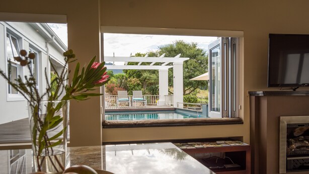 A living room with a view of a pool, a fireplace, and a TV mounted on the wall. - Modern house, solar-heated pool & stunning views, at Kingswood Golf Resort (George)