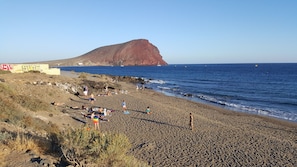 Ein Strand mit badenden Personen und Spaziergängern, felsigem Gelände und einer großen Anhöhe im Hintergrund.