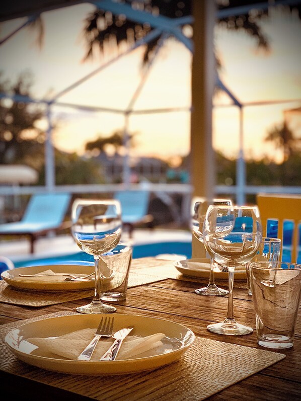 A dining table set with plates, cutlery, and glasses, overlooking a pool area with lounge chairs and palm trees.