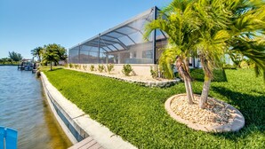 A modern house with a large glass window, a well-maintained lawn, and a palm tree. - SW Cape Coral Waterfront Villa "Summerset" | Pool + Jacuzzi | south-facing | (Cape Coral)