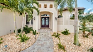 A house with a stone pathway leading to a dark door, surrounded by palm trees and a gravel garden. - SW Cape Coral Waterfront Villa "Summerset" | Pool + Jacuzzi | south-facing | (Cape Coral)