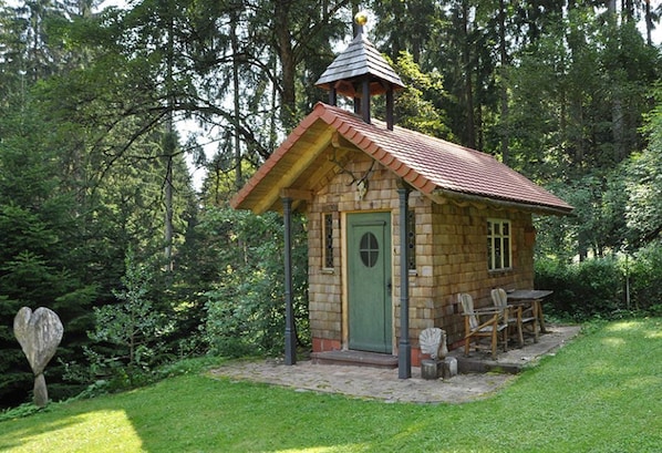 Una pequeña cabaña de piedra con puerta verde, techo de tejas y un porche con sillas de madera.