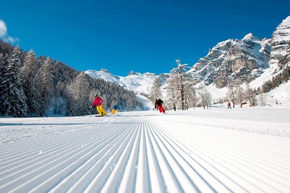 Ein verschneiter Skipisten mit Skifahrern, Bäumen und Bergen im Hintergrund.