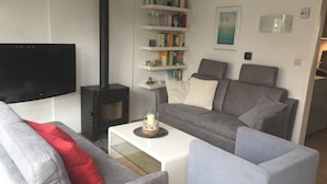 A modern living room with a flat-screen TV, a black wood-burning stove, grey sofas, a white coffee table, and a bookshelf with various items. - Corner house 3 bedrooms on the Grevelingen Sea in the park Aquadelta, free Wi-Fi (Bruinisse)