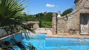 Una piscina con mosaicos, paredes de piedra y vistas a una colina con una torre.