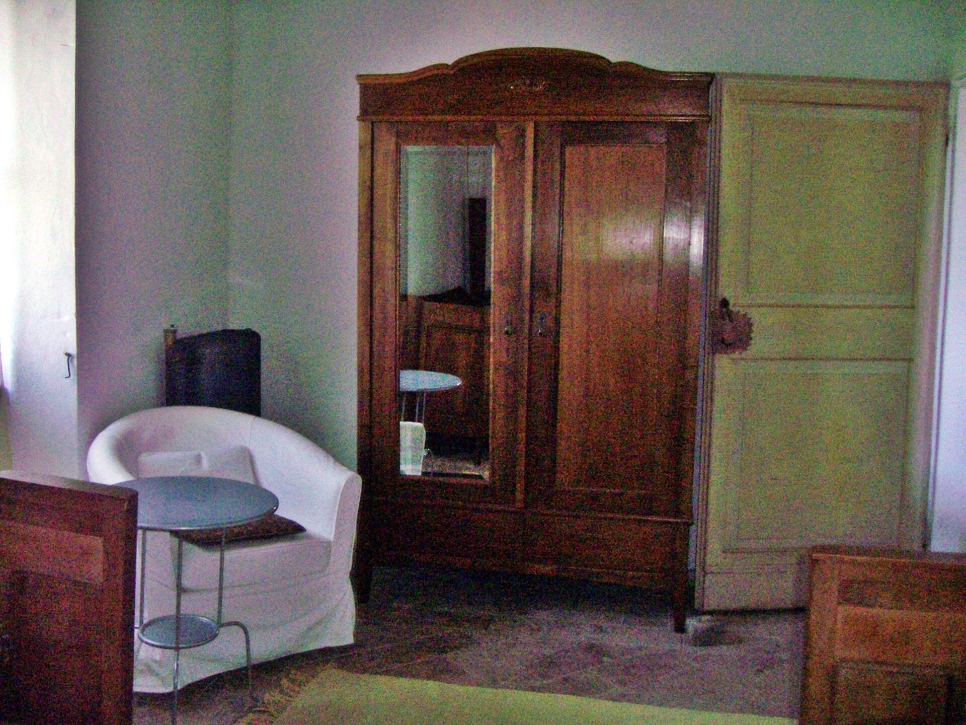 A room with a white armchair, a wooden wardrobe, a small table, and a door.