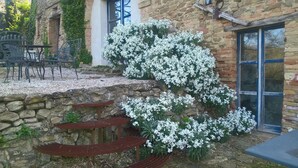 Un muro de piedra con un banco de metal, plantas florecientes blancas y un edificio con una ventana y una puerta.