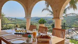 Ein Frühstückstisch mit schöner Aussicht, gedeckt mit Tischdecke, Tellern, Besteck, Gläsern und einem Obstkorb.