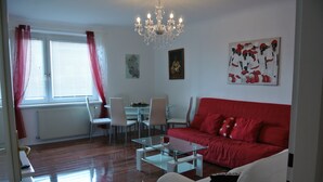 A living room with a red sofa, white armchairs, a glass coffee table, and a chandelier. There is a dining area with a table and chairs, and a window with curtains. - Totally new apartment, central location, competitive price, WiFi, elevator (Wien)