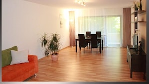 Una sala de estar con piso de madera, una mesa de comedor con sillas, un sofá con cojines, una planta en maceta y una lámpara.