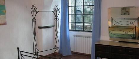 Un dormitori amb estructura de llit metàl·lica, una finestra amb cortines blaves, un radiador i una petita taula de nit.