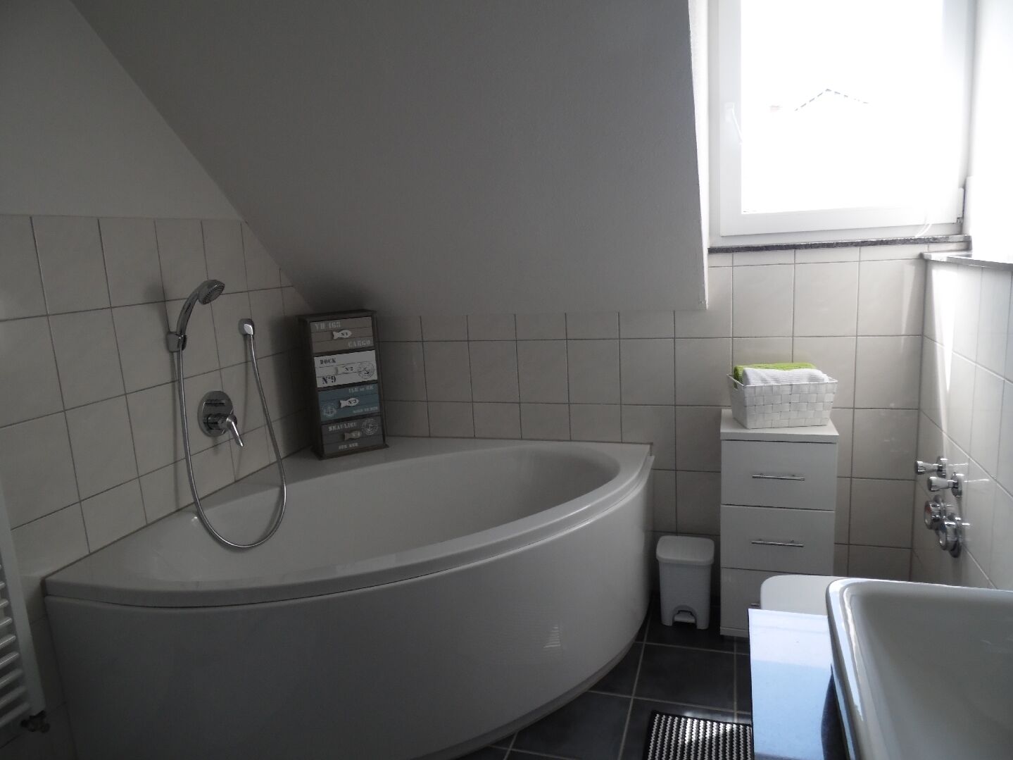Une salle de bain avec une grande baignoire, des toilettes et un petit lavabo. Il y a une fenêtre qui laisse entrer la lumière naturelle et un mur recouvert de carreaux.