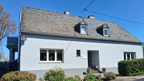 A two-story building with a grey roof, a small porch, and a sign indicating a business. - Apartment at St. James, amidst the nature park Südeifel, on the edge of Islek (Lascheid)