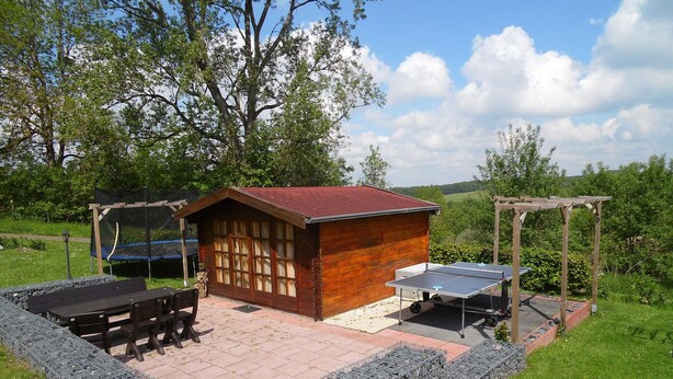 A wooden cabin with a red roof, a trampoline, and a ping pong table in an outdoor setting. - Apartment at St. James, amidst the nature park Südeifel, on the edge of Islek (Lascheid)