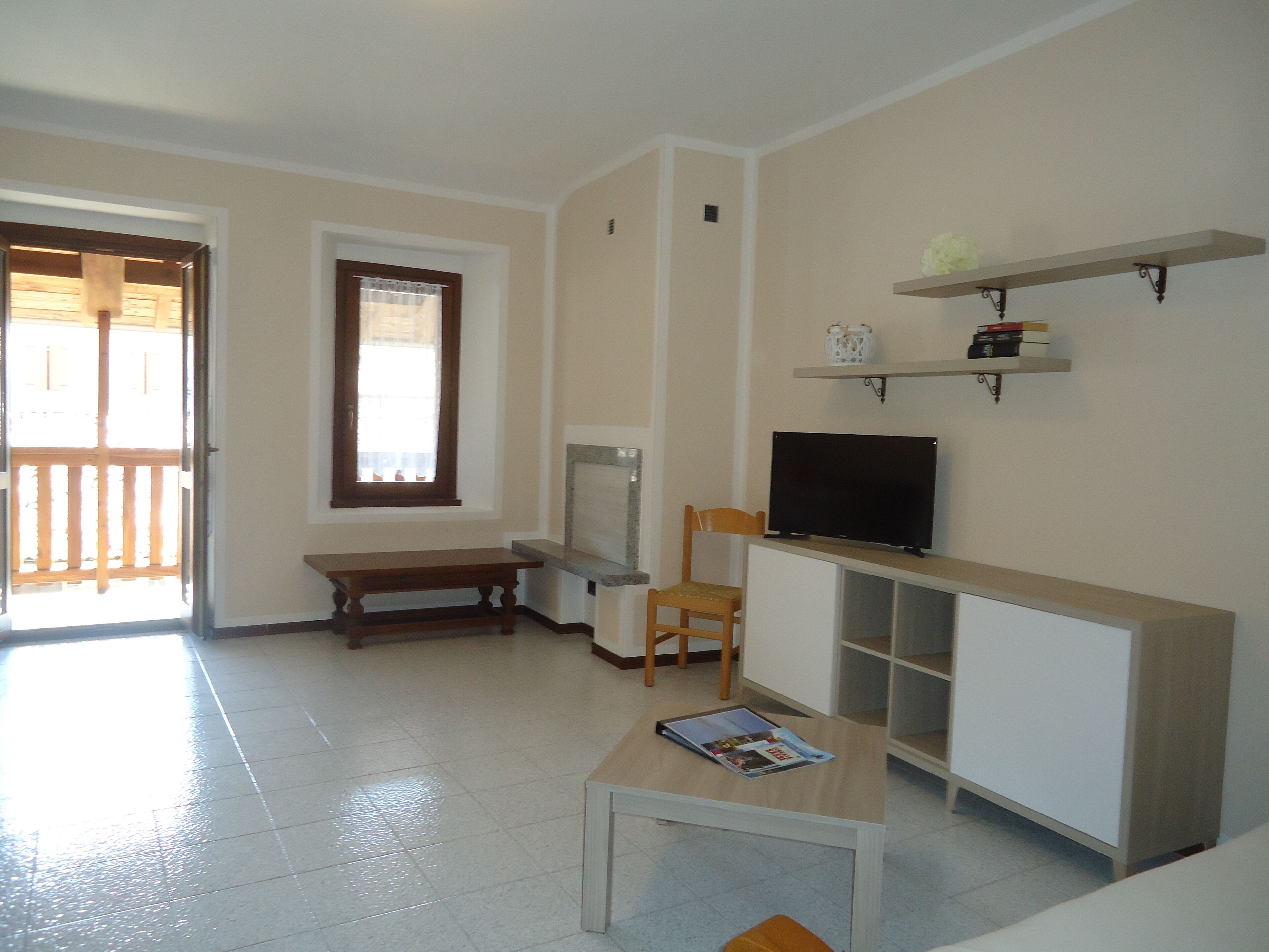 A living room with a television, a bench, a table with a magazine, a window, and a door leading outside.