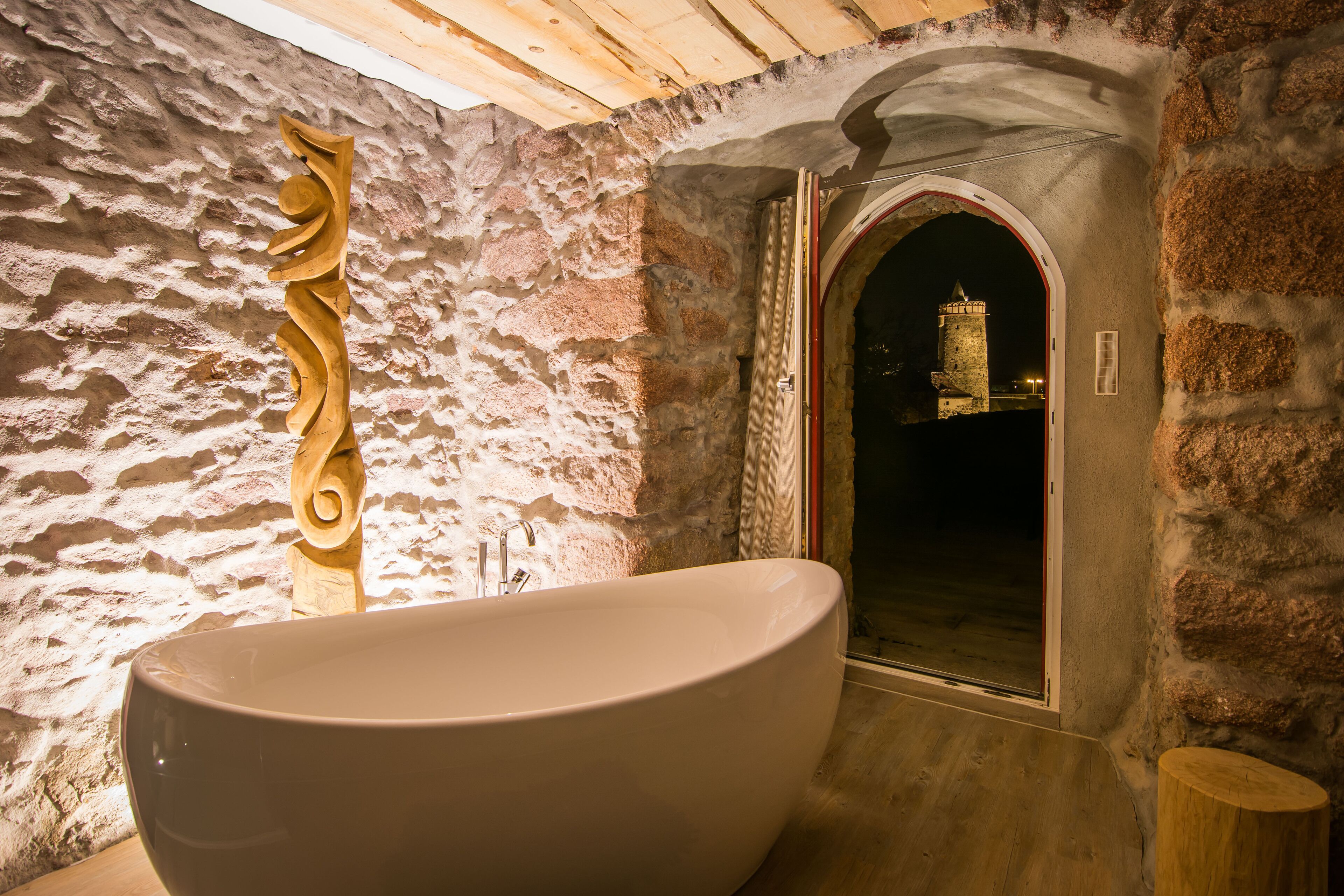 A large freestanding bathtub in a stone-walled bathroom with a wooden sculpture and a view of a tower through an arched doorway.
