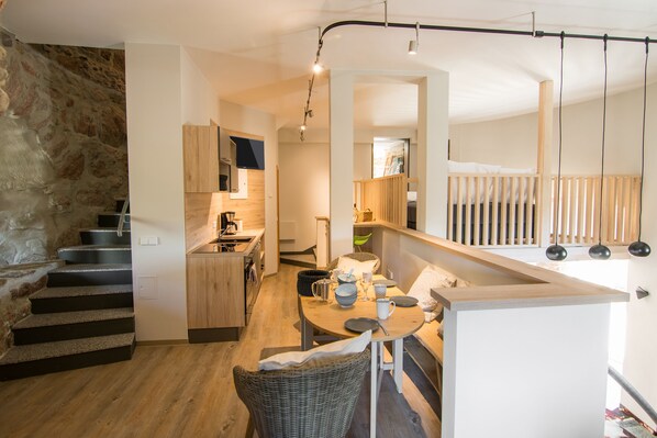 Una cocina moderna con armarios de madera, un comedor con mesa y sillas, y una escalera con un muro de piedra a un lado.