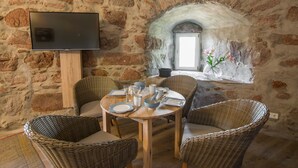 Ein Zimmer mit Steinwänden, ein runder Holztisch gedeckt für das Essen, Korbstühle, ein Fernseher auf einem Schrank und ein Fenster mit Blumen.