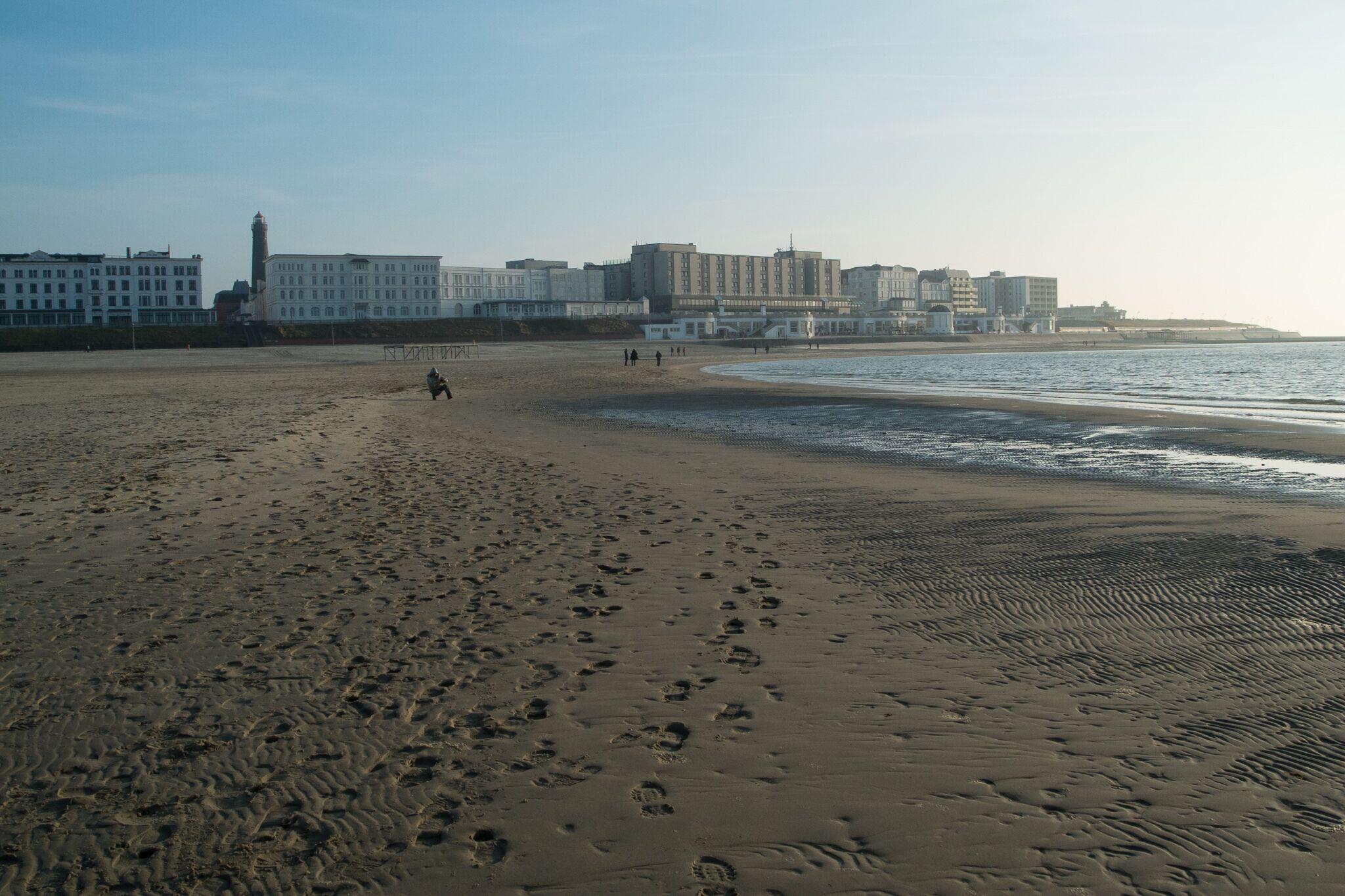 A wide sandy beach with footprints, a lighthouse, and buildings in the background.