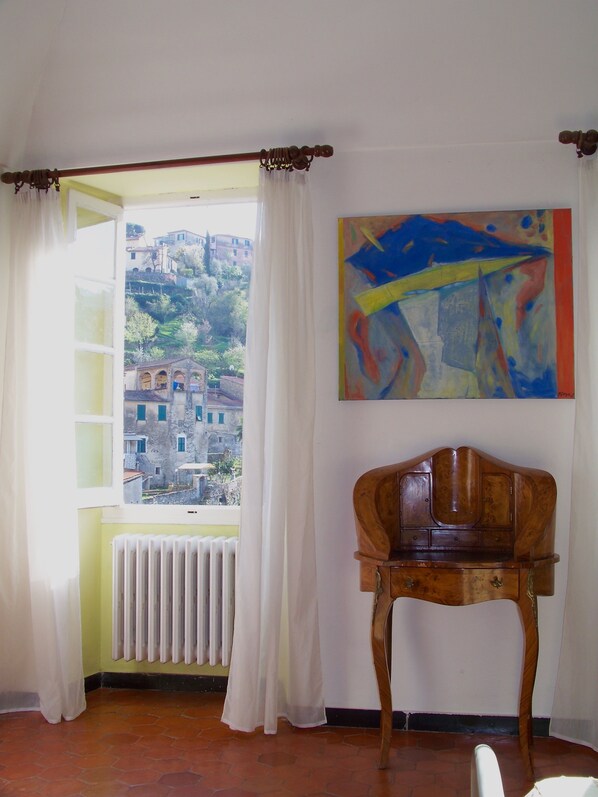 Una stanza con vista sui palazzi attraverso una finestra, un termosifone, una sedia di legno e un quadro appeso al muro.