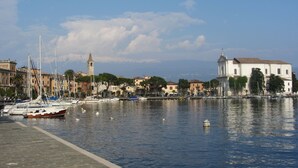 A waterfront view with boats docked, a prominent tower, and a white building with a bell tower. - Apartment with air conditioning, sea view, swimming pool, Wi-Fi, quiet hill, parking. (Toscolano Maderno)