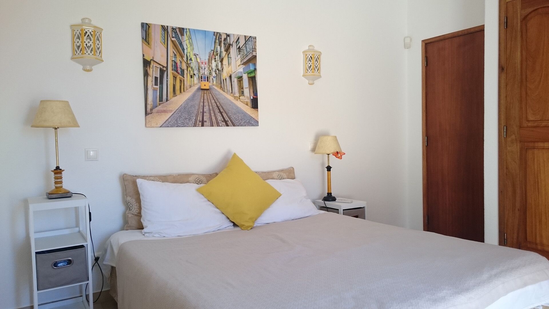Une chambre à coucher avec un lit, deux tables de chevet avec des lampes, une porte en bois et un tableau représentant une scène de rue, accroché au mur.