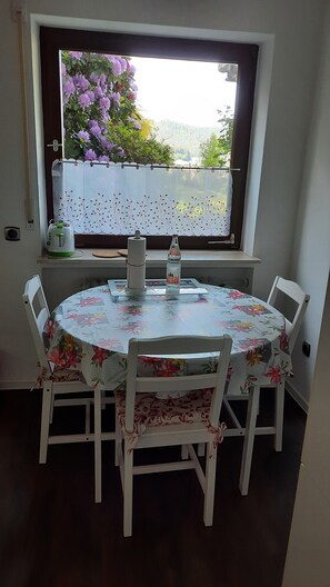 A dining area with a round table, floral tablecloth, white chairs, a window with a view of flowers and greenery, and a small shelf with a kettle and a bottle.