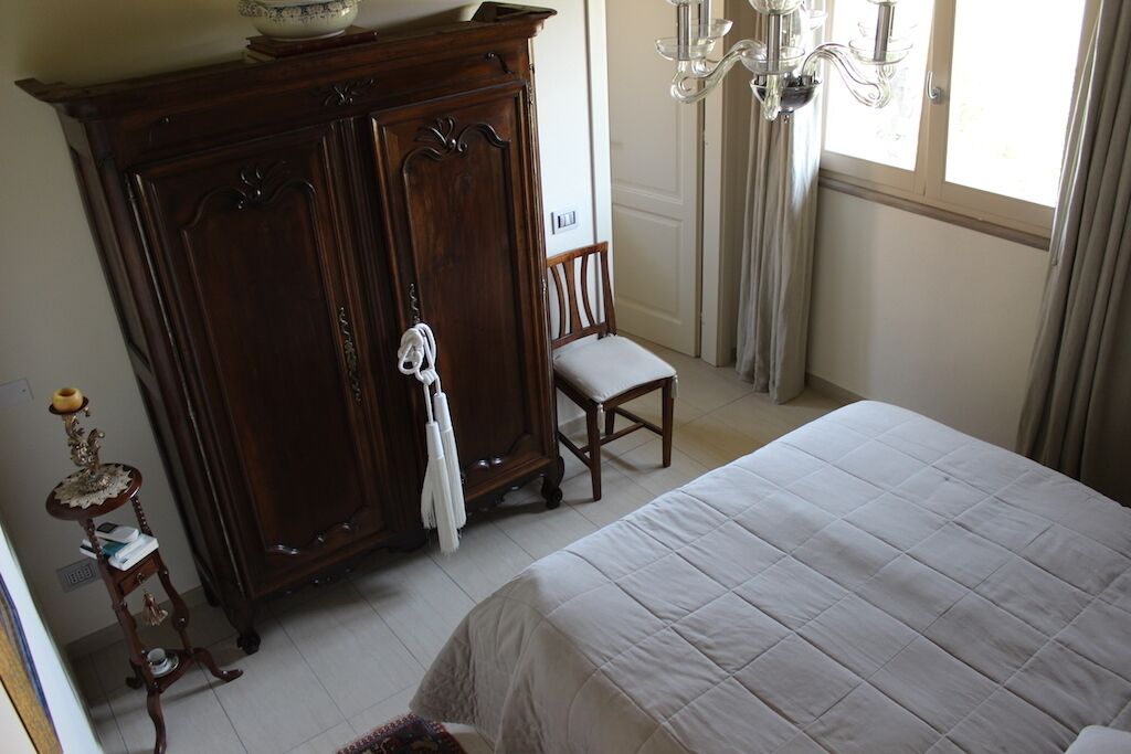 A bedroom with a large bed, a wooden wardrobe, a chair, and a small table with a lamp.