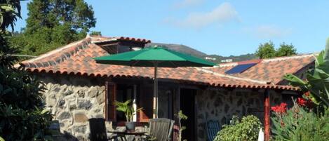 A stone building with a tiled roof, a patio with a table and chairs, and a green umbrella.