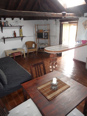 Quarto acolhedor com uma mesa de jantar de madeira, um sofá e uma cadeira. Há uma estante com livros e um objeto decorativo.
