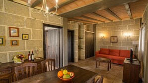 Un coin repas rustique avec une table en bois, des chaises et un panier de fruits. Le salon comprend un canapé rouge et un téléviseur. Les murs sont en pierre et des tableaux encadrés y sont accrochés.