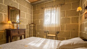 Une chambre avec un mur en pierre, une commode en bois, une table de chevet avec une lampe, une fenêtre avec un rideau léger et un lit recouvert d’une couvre-lit à motifs.