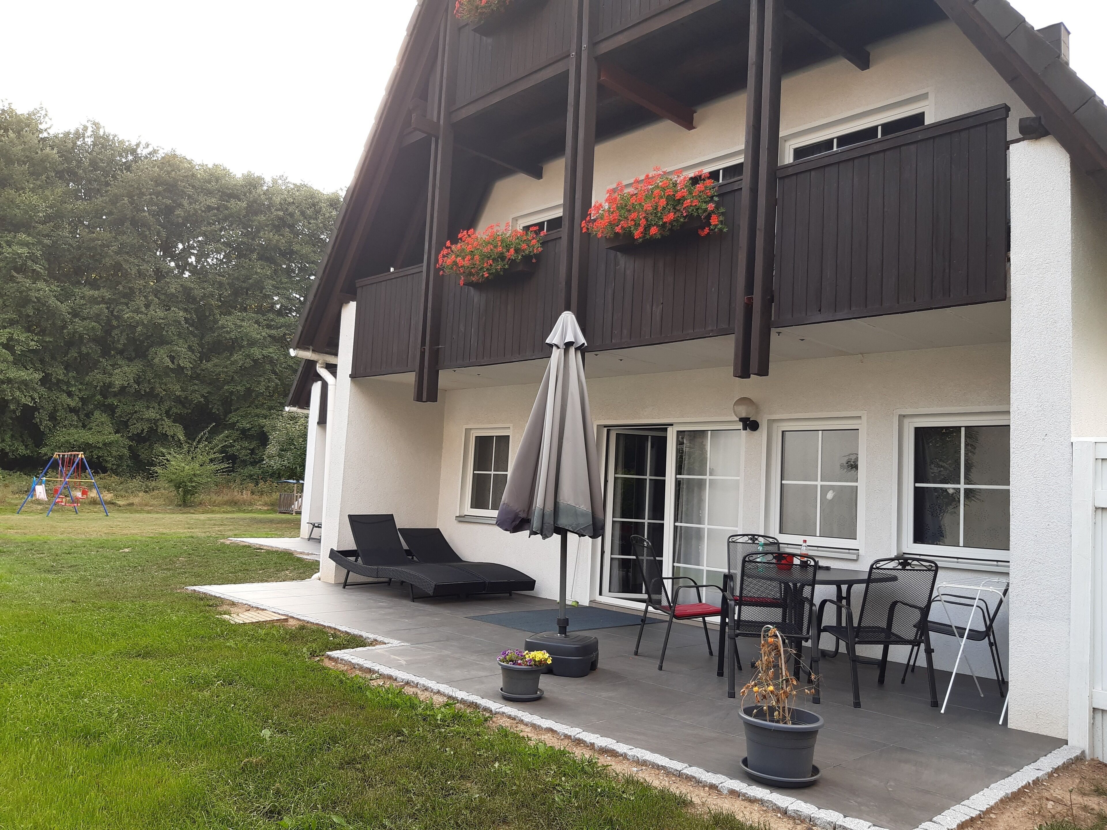 Ett tvåvåningshus med en balkong, en utomhusplats med sittmöbler och en gunga i trädgården.