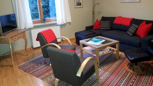 Un salón con sofá, sillones, una mesa de centro, televisión y una ventana con cortinas.